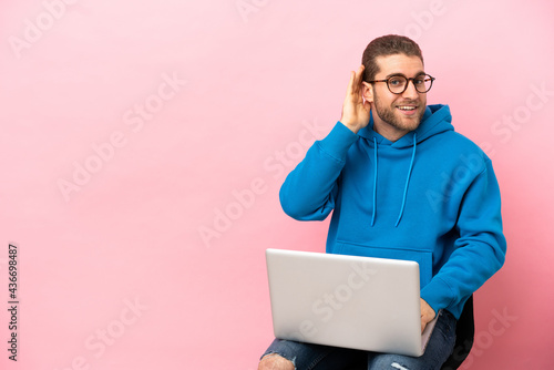 Young man sitting on a chair with laptop listening to something by putting hand on the ear