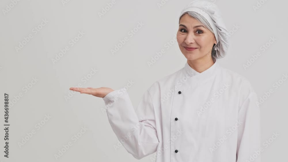A smiling chef cook woman is standing in a confident pose while presenting something isolated over grey wall in the studio