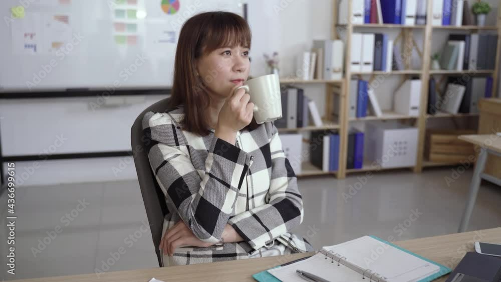 pretty asian businesswoman is looking into distance and thinking with a satisfied smile while enjoying tea at desk in the bright workplace. pretty asian businesswoman is looking into distance and thinking with a satisfied smile while enjoying tea at desk in the bright workplace.