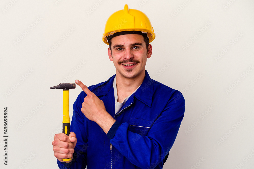 Young caucasian worker man holding a hammer isolated on white background smiling and pointing aside, showing something at blank space.