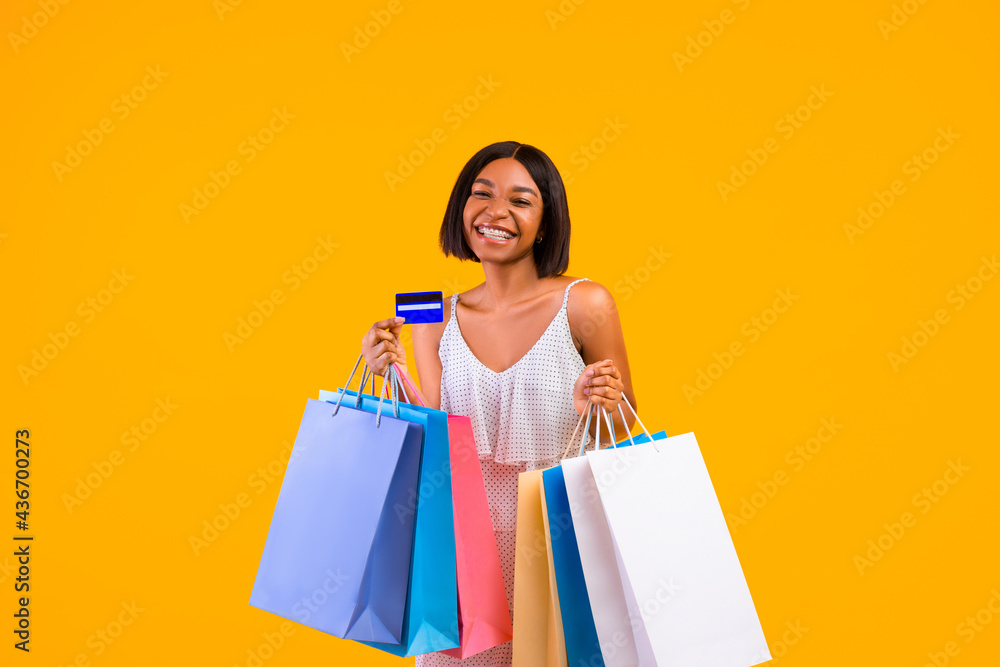 Seasonal shopping. Smiling black woman holding gift bags and credit card, smiling at camera on orange studio background Seasonal shopping. Smiling black woman holding gift bags and credit card, smiling at camera on orange studio background