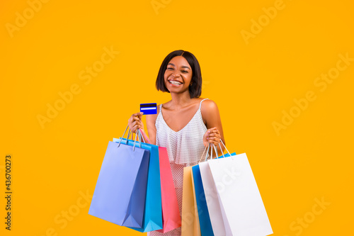 Seasonal shopping. Smiling black woman holding gift bags and credit card, smiling at camera on orange studio background