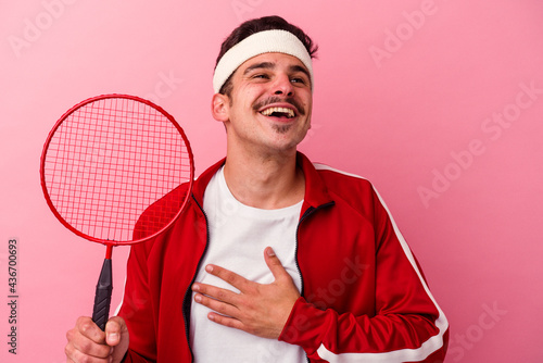 Young caucasian man playing badminton isolated on pink background laughs out loudly keeping hand on chest. © Asier