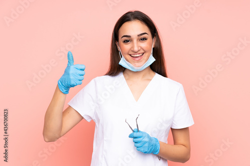 Woman dentist holding tools isolated on pink background giving a thumbs up gesture
