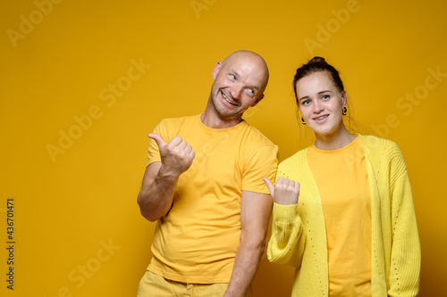 Cute, benevolent man and woman in yellow clothes invite somewhere by pointing their thumb to the side. Copy space. Bright background.