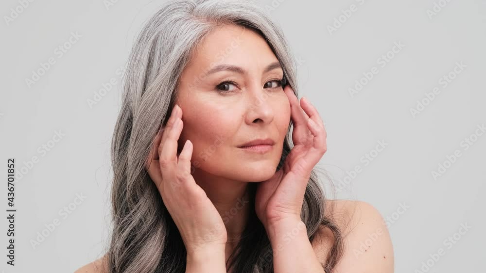 A pleased elder woman is looking at her face standing isolated over grey wall in the studio A pleased elder woman is looking at her face standing isolated over grey wall in the studio