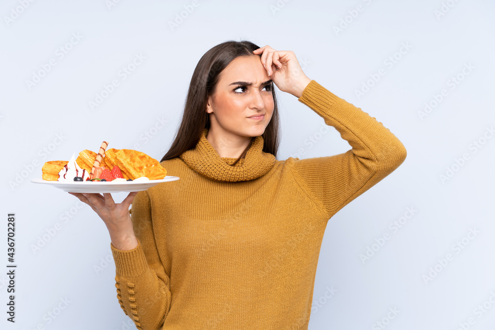 Young caucasian woman holding waffles isolated on blue background having doubts and with confuse face expression Young caucasian woman holding waffles isolated on blue background having doubts and with confuse face expression