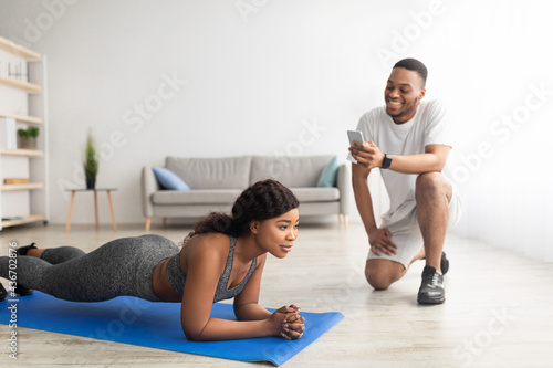 Millennial black couple training as team at home, athletic woman standing in plank pose, her boyfriend noting time