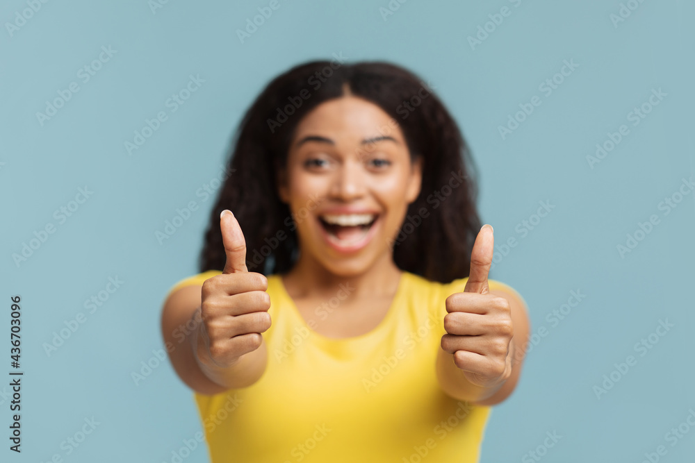 Joyful african american woman showing thumbs up with both hands and smiling to camera, blue background, selective focus Joyful african american woman showing thumbs up with both hands and smiling to camera, blue background, selective focus