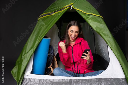 Young caucasian woman inside a camping green tent with phone in victory position
