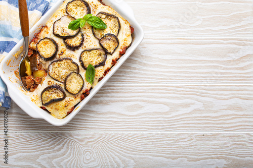 Greek mediterranean dish Moussaka with baked eggplants, ground beef in white ceramic casserole with napkin on wooden white background from above, traditional dish of Greece, space for text