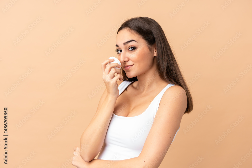 Fototapeta premium Young caucasian woman isolated on beige background with moisturizer and smelling it