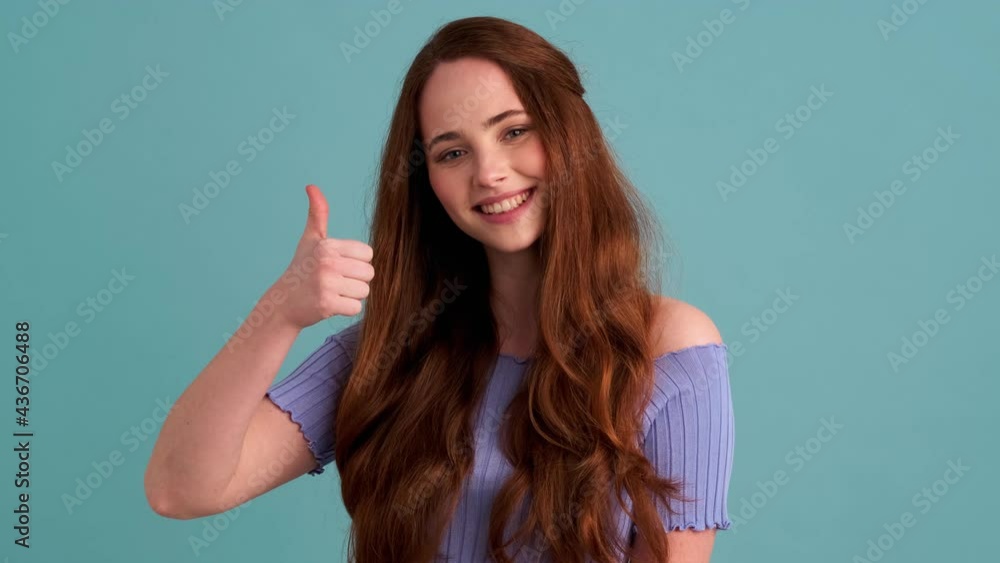 A pleased woman is showing thumb-up gesture standing isolated over blue wall in the studio A pleased woman is showing thumb-up gesture standing isolated over blue wall in the studio