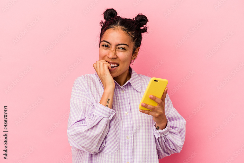 Young latin woman holding a mobile phone isolated on pink background biting fingernails, nervous and very anxious. Young latin woman holding a mobile phone isolated on pink background biting fingernails, nervous and very anxious.