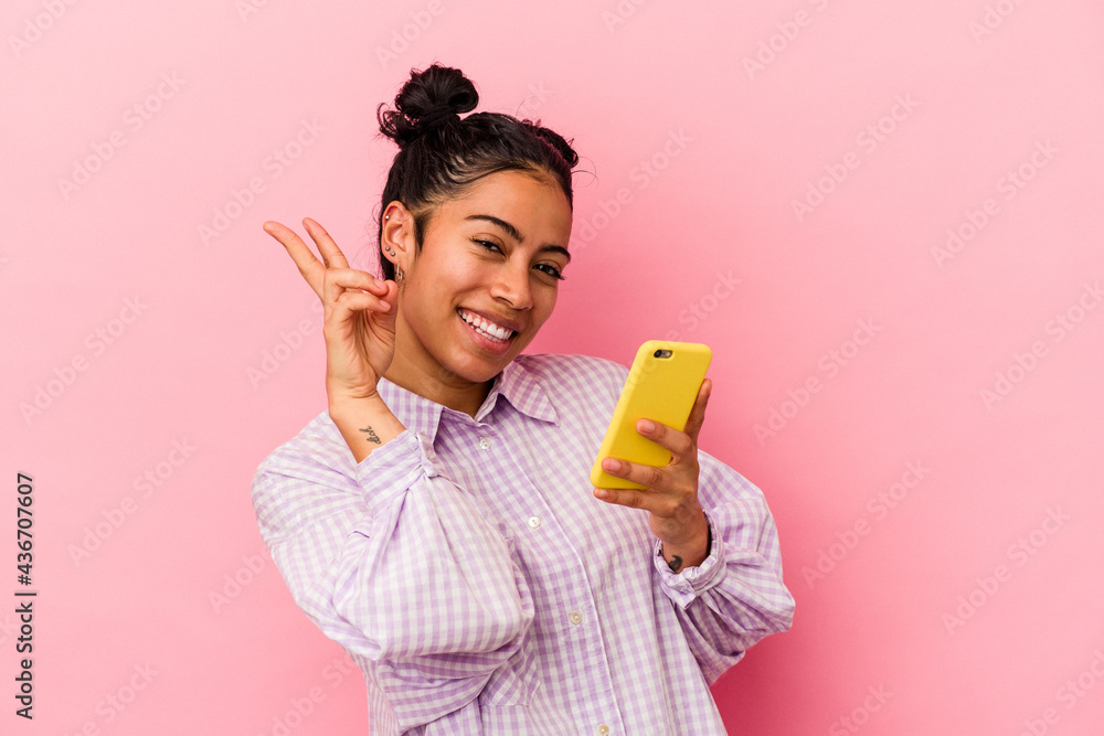 Young latin woman holding a mobile phone isolated on pink background joyful and carefree showing a peace symbol with fingers.