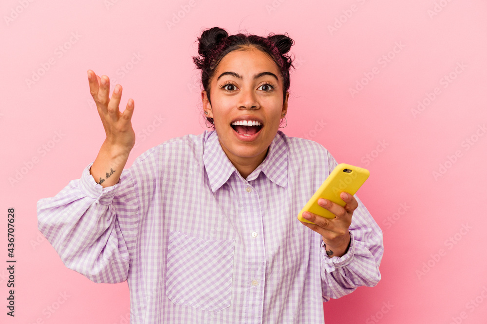 Young latin woman holding a mobile phone isolated on pink background receiving a pleasant surprise, excited and raising hands. Young latin woman holding a mobile phone isolated on pink background receiving a pleasant surprise, excited and raising hands.