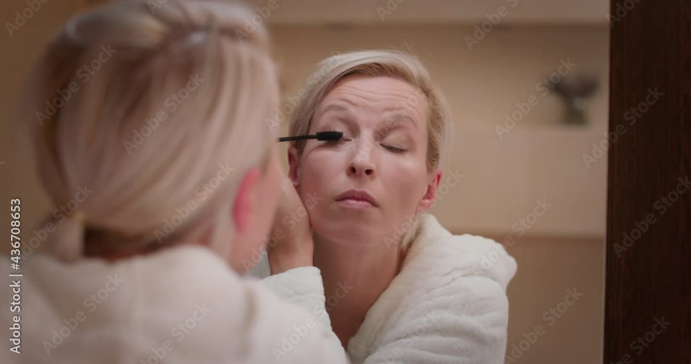 Woman looking in the mirror in the bathroom, applies mascara on her eye lashes Woman looking in the mirror in the bathroom, applies mascara on her eye lashes