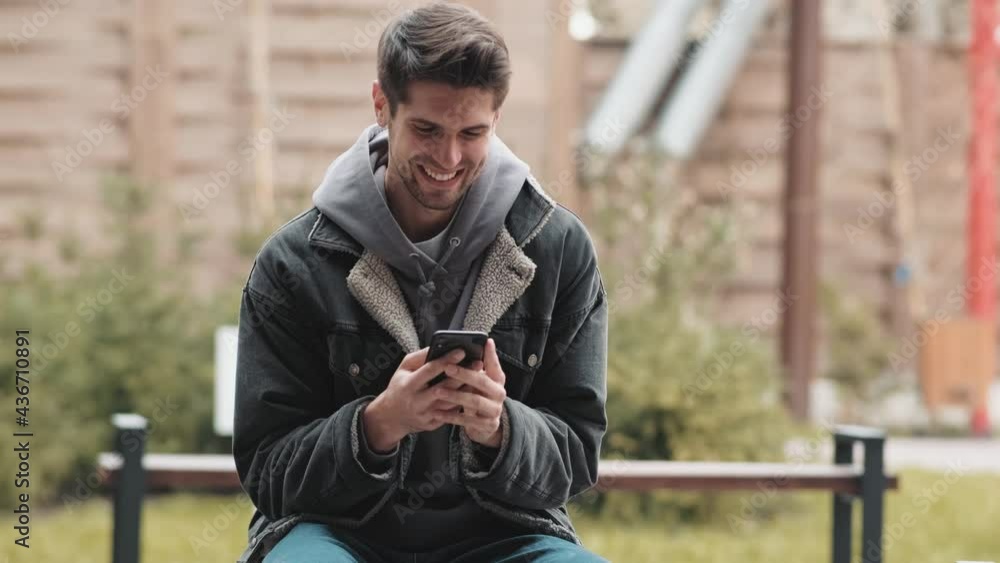 A smiling man in a hoodie and denim jacket looking at the phone while sitting on a bench in the street A smiling man in a hoodie and denim jacket looking at the phone while sitting on a bench in the street