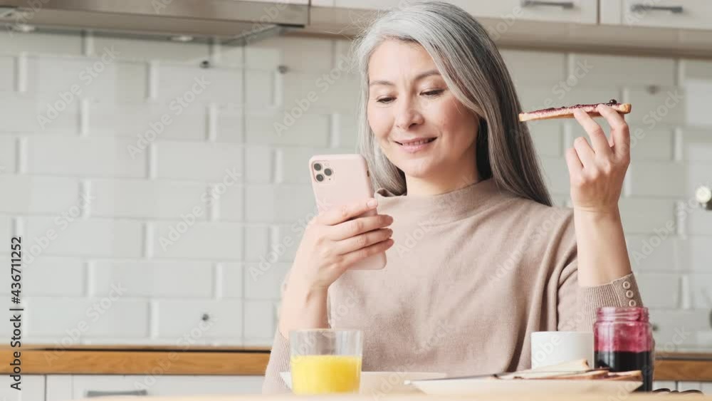 A positive elder woman is eating toasts with jam while using her smartphone sitting in the kitchen at home A positive elder woman is eating toasts with jam while using her smartphone sitting in the kitchen at home