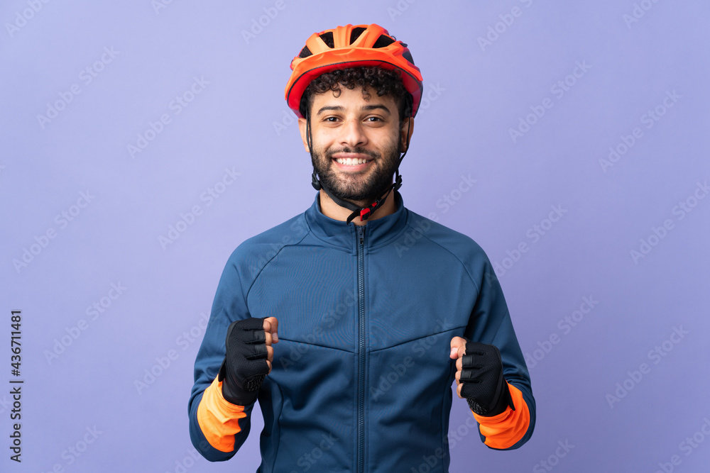 Young Moroccan cyclist man isolated on purple background celebrating a victory in winner position Young Moroccan cyclist man isolated on purple background celebrating a victory in winner position
