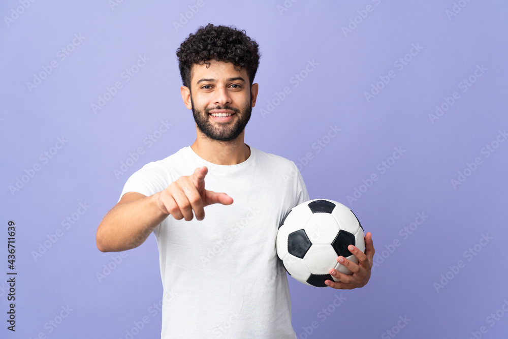 Young Moroccan man isolated on purple background with soccer ball and pointing to the front Young Moroccan man isolated on purple background with soccer ball and pointing to the front