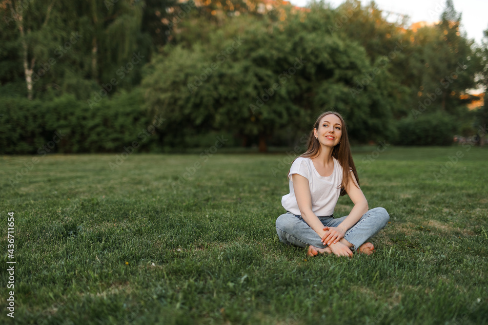 A Caucasian girl with brown hair in blue jeans and a white T-shirt is sitting on the grass . Sunset. A Caucasian girl with brown hair in blue jeans and a white T-shirt is sitting on the grass . Sunset.