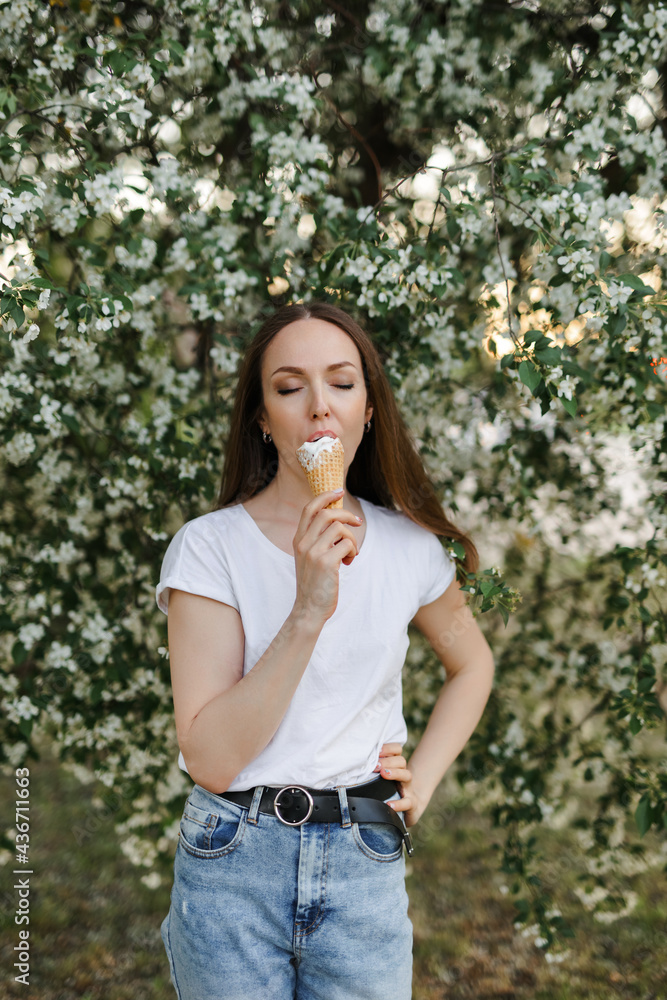 Caucasian girl with brown hair in blue jeans and a white T-shirt eats ice cream. The concept of spring flowering. Apple tree in bloom. Caucasian girl with brown hair in blue jeans and a white T-shirt eats ice cream. The concept of spring flowering. Apple tree in bloom.