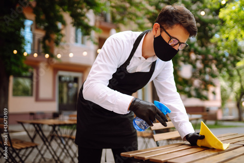 Waiter in protective face mask and gloves disinfecting tables at outdoor cafe due to coronavirus epidemic. COVID-19.
