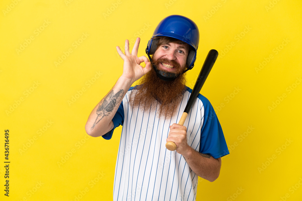 Redhead baseball player man with helmet and bat isolated on yellow background showing ok sign with fingers