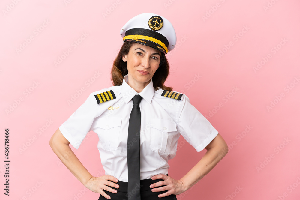 Fototapeta premium Airplane middle aged pilot woman isolated on pink background posing with arms at hip and smiling