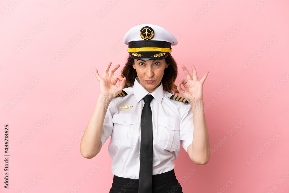 Airplane middle aged pilot woman isolated on pink background showing an ok sign with fingers Airplane middle aged pilot woman isolated on pink background showing an ok sign with fingers