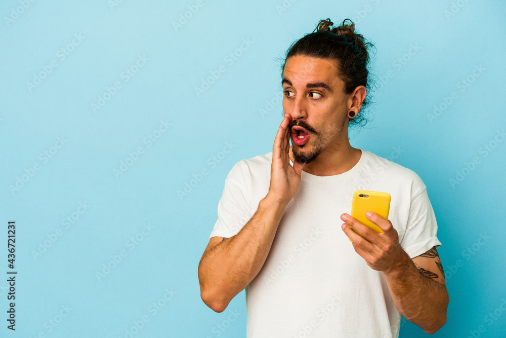 Young caucasian man with long hair holding mobile phone isolated on blue background is saying a secret hot braking news and looking aside Young caucasian man with long hair holding mobile phone isolated on blue background is saying a secret hot braking news and looking aside