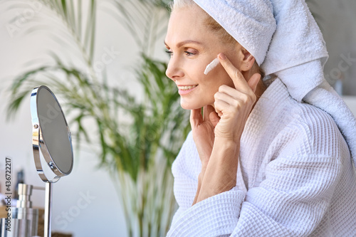 Gorgeous senior older adult 50 years old blonde woman wearing bathrobe and turban towel in bathroom applying moisturizing tightening face skin treatment, looking at mirror. Morning beauty routine. photo