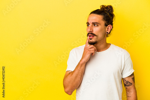 Young caucasian man with long hair isolated on yellow background looking sideways with doubtful and skeptical expression.