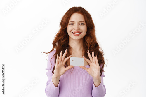 Smiling cute girl with ginger hair shows her new bank credit card, shopping in stores contactless, buying things with deposit money, standing over white background
