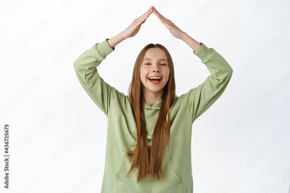 Smiling happy girl shows home roof gesture with hands above head, laughing and looking upbeat, making rooftop sign, standing in hoodie against white background Smiling happy girl shows home roof gesture with hands above head, laughing and looking upbeat, making rooftop sign, standing in hoodie against white background
