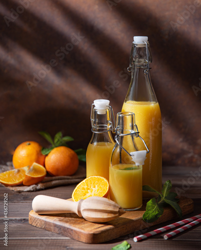 Fresh homemade orange juice in three bottles with citrus fruits on cutting board with morning shadows on a brown wooden background. Front view and copy space