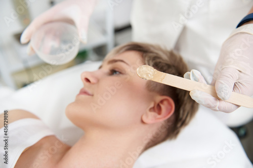 Appliance of anaesthetic gel on woman's face for making the laser removal hair procedure non painful. Professional laser treatment in modern SPA clinic. Focus on wooden spatula