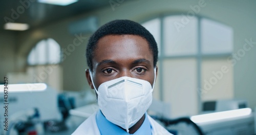 Close up shot of handsome African American young male doctor with protective mask on face looking at camera at workplace in clinic lab. Male doctor in laboratory, hospital, coronavirus pandemic