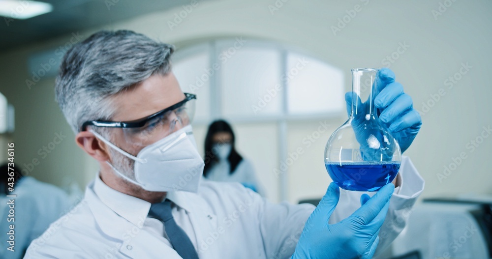 Caucasian senior male pharmacist in mask and goggles working in lab looking at blue liquid in flask doing medical research, providing experiment in laboratory, science, close up concept, healthcare Caucasian senior male pharmacist in mask and goggles working in lab looking at blue liquid in flask doing medical research, providing experiment in laboratory, science, close up concept, healthcare