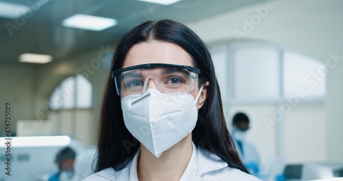 Close up portrait of happy young beautiful Caucasian female medical specialist wearing protective mask and goggles and looking at camera. Healthcare worker, lab scientist, laboratory work