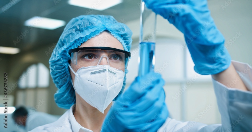 Close up of Caucasian pretty young female medical scientist holding glass test tube with blue liquid in laboratory. Medic experiment to detect virus chemist working on coronavirus vaccine cure concept Close up of Caucasian pretty young female medical scientist holding glass test tube with blue liquid in laboratory. Medic experiment to detect virus chemist working on coronavirus vaccine cure concept