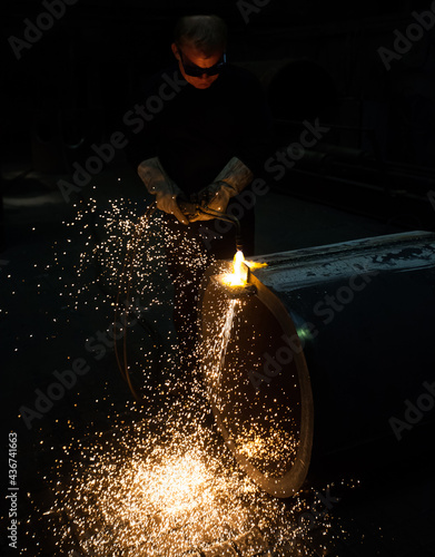 Welder worker in protective glasses cuts metal pipe with gas torch. Dark tones, bright yellow flame and sparks.