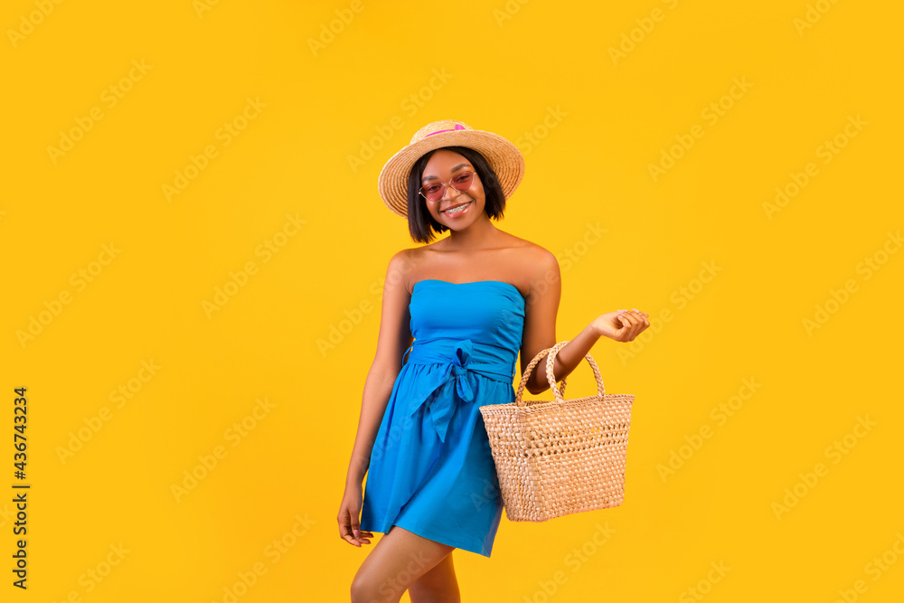 Portrait of young pretty black lady in straw hat and blue dress smiling, holding trendy beach bag over orange background