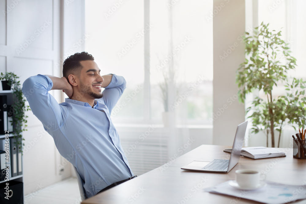 Confident Arab company employee leaning on chair with hands behind head, relaxing after completed job at workplace Confident Arab company employee leaning on chair with hands behind head, relaxing after completed job at workplace