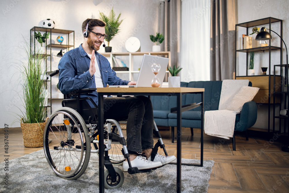 Bearded man in eyeglasses and headset talking and gesturing during video conference on modern laptop. Freelancer with disabilities having opportunity to work from home. Bearded man in eyeglasses and headset talking and gesturing during video conference on modern laptop. Freelancer with disabilities having opportunity to work from home.