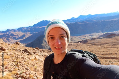 A cheerful young woman with hazel eyes wearing an ice cap taking a selfie on top of Moses mountain during the sunrise in Saint Catherine in Egypt