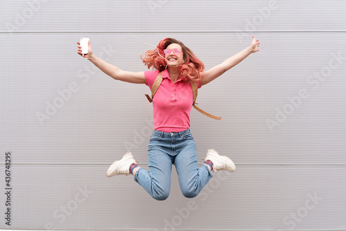 Fototapeta Naklejka Na Ścianę i Meble -  Fashion portrait of pretty smiling hipster woman in sunglasses and colored red hairs with smartphone against the colorful grey wall. Jumping
