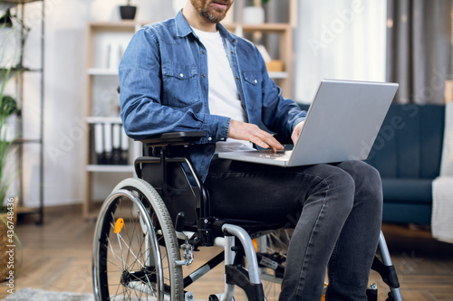 Close up of handicapped young man working on wireless laptop while sitting in wheelchair at living room. Remote work for people with disabilities.