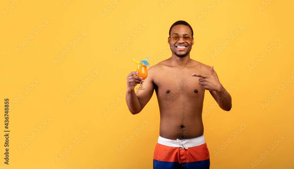 Joyful black guy with bare torso having beach vacation, pointing at tropical cocktail on yellow background, free space